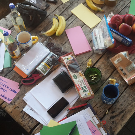 Vogelperspektive, Halbtotale: Tischfläche aus altem Holz mit Snacks und Arbeitsmaterial für ein Seminar. Zwei Personen sitzen am Tisch und arbeiten mit Moderationskarten.